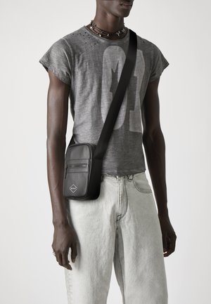 Geantă neagră din piele, tip crossbody, cu buzunar cu fermoar, formă rectangulară și curea ajustabilă, purtată peste un tricou gri cu grafică și blugi deschiși.