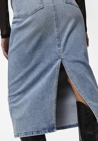 Jupe en denim bleu clair avec une fente arrière, comportant deux poches arrière et un ourlet effiloché, associée à une botte noire.