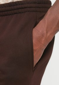 Mano derecha descansando dentro del bolsillo de unos pantalones de cintura elástica marrón oscuro, mostrando la textura de la tela y detalles de la piel.