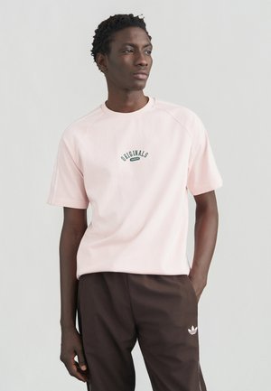 Lyserød T-shirt med kortærmer og tekstur, med "ORIGINALS" logo i grøn, båret med mørkebrune bukser og tre-stribede accenter.