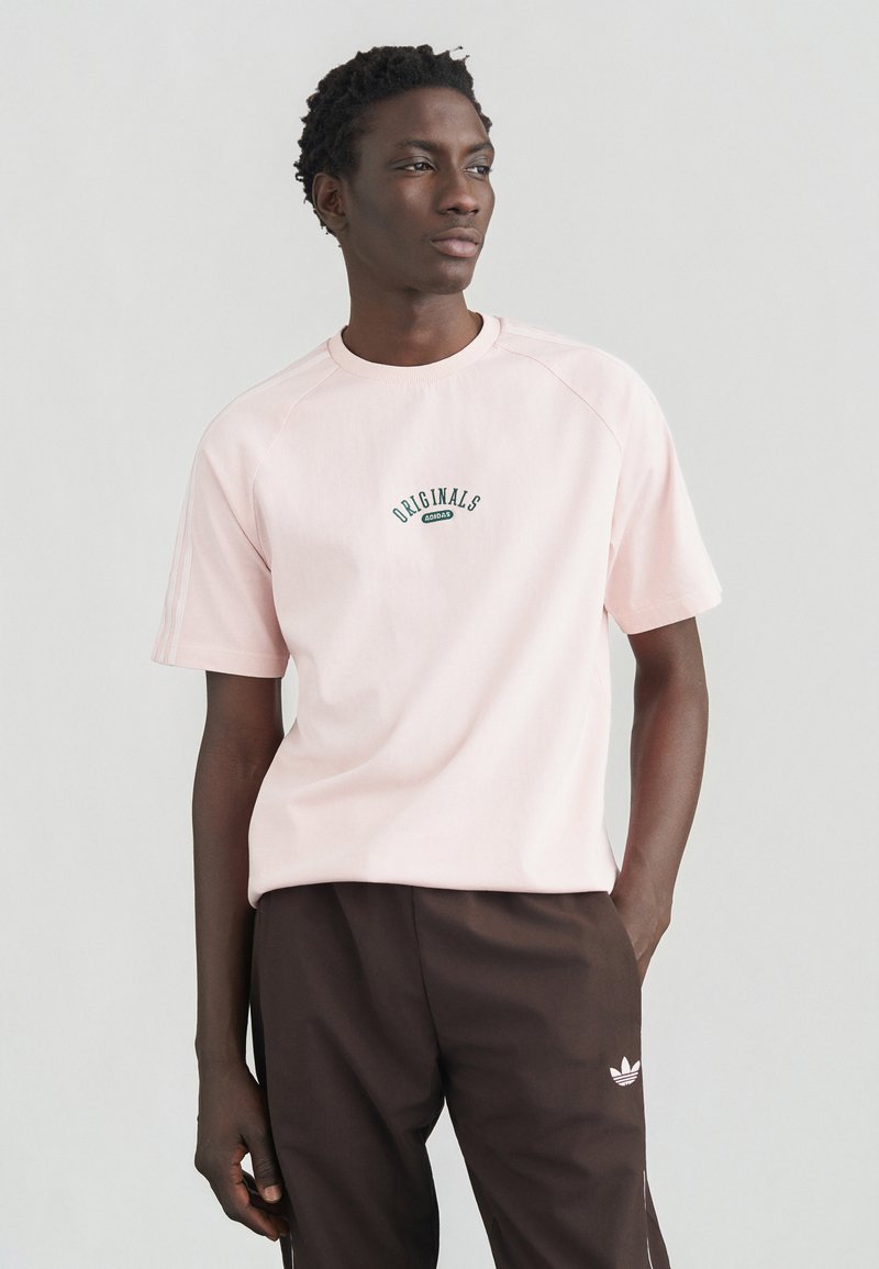 Ροζ ανοιχτό ριμπ κοντομάνικο μπλουζάκι με υφή, με το λογότυπο "ORIGINALS" σε πράσινο, φορεμένο με σκούρο καφέ παντελόνι και λεπτομέρειες με τρεις ρίγες.