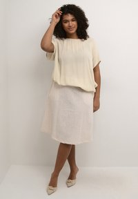 Light, flowy cream top with short sleeves paired with a soft, light beige knee-length skirt; model wearing neutral-colored, pointed flats.