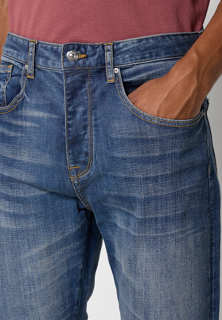 Primo piano di una persona che indossa jeans in denim blu con la mano nella tasca destra e una camicia bordeaux infilata dentro.