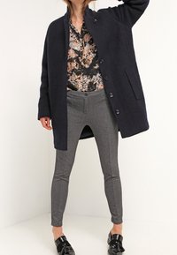 Manteau en laine marine avec fermeture à boutons et poches latérales, porté avec un pantalon gris ajusté et des chaussures noires ornées d'accents décoratifs.