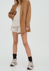 Blazer marron oversize porté sur un haut blanc texturé et un short, associé à des baskets épaisses et des chaussettes noires côtelées. Palette de couleurs neutres.