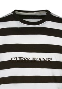 T-shirt rayé noir et blanc avec un col rond, arborant le texte brodé "GUESS JEANS" en marron sur le devant. Texture en tissu coton.