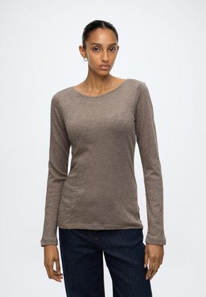 Femme aux cheveux foncés tirés en arrière, portant un haut taupe à manches longues et un jean bleu foncé sur un fond clair uni.