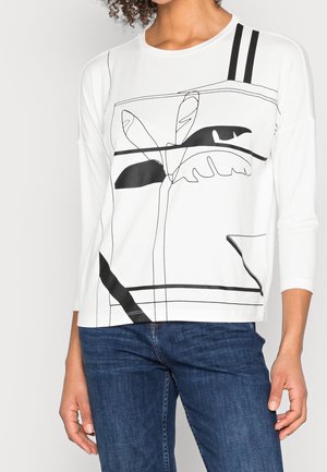 Kvinde iført hvid langærmet top med abstrakt sort plante-design og to lodrette striber, kombineret med blå jeans, stående i afslappet positur.