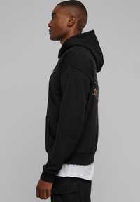 Fekete kapucnis pulóver pamutkeverék anyagból, első zsebbel, bordázott mandzsettákkal és szegéllyel. Hátul grafikai nyomat található. Normál szabású, állítható kapucnival.