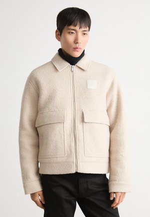 Beige fleecejakke med krave, lynlås foran, to frontlommer og et firkantet logo-patch på brystet. Båret over en sort turtleneck.
