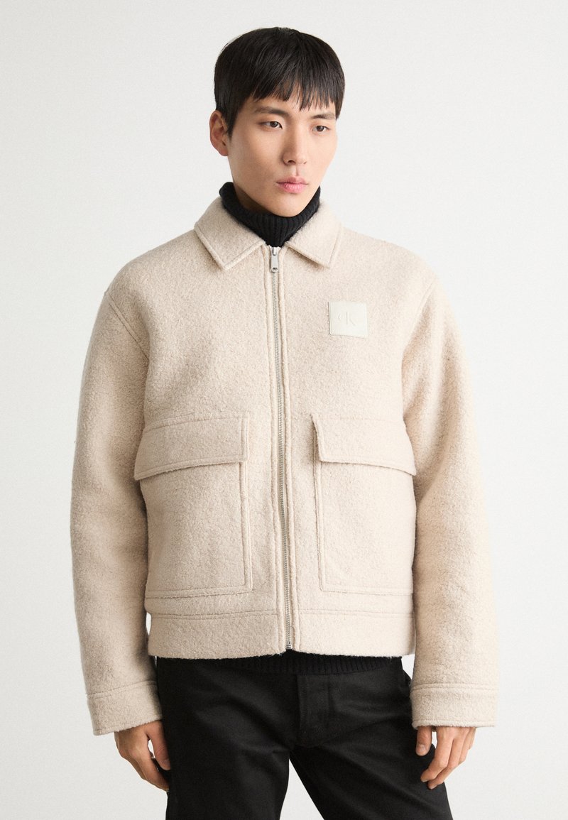 Beige fleecejacka med krage, dragkedja framtill, två framfickor och en fyrkantig logopatch på bröstet. Buren över en svart polotröja.
