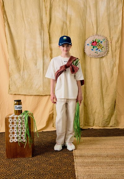 Jeune personne vêtue d'une tenue blanche et d'une casquette bleue tenant des haricots verts longs, debout près d'un bocal étiqueté « Cornichons » posé sur une caisse en bois avec un tissu crocheté.