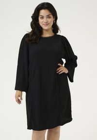 Vestido negro con mangas largas abullonadas, tejido texturizado, escote redondo y detalles en forma de onda a lo largo de los lados, diseño hasta la rodilla.