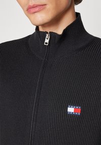 Top zipato a coste nera con colletto alto e una piccola patch colorata del logo Tommy Jeans sul petto. Dettaglio in tessuto testurizzato visibile.