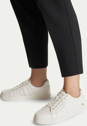 Osoba ubrana w czarne, krótkie spodnie i białe pikowane trampki z złotymi przelotkami i akcentami, stojąca na jasnym tle.