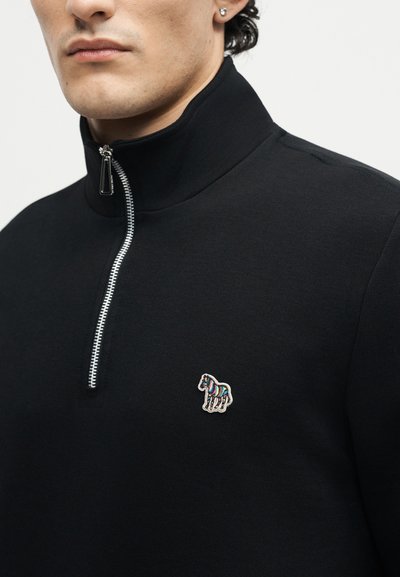 Άνδρας που φοράει μαύρο πουλόβερ με φερμουάρ και υψηλό γιακά, με μικρό πολύχρωμο λογότυπο ζέβρας στο αριστερό στήθος.