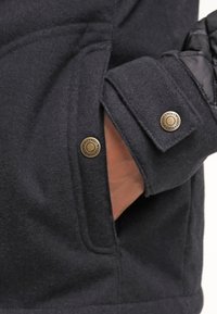 Veste bleu marine en tissu texturé, avec poignets boutonnés et détails matelassés. Poche apparente et finitions en laiton ajoutent une touche fonctionnelle.