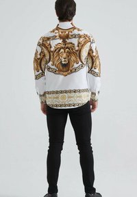 Un homme debout de dos, portant une chemise blanche ornée d'une grande tête de lion brune détaillée et de motifs de chaînes dorées sur le dos et les manches.