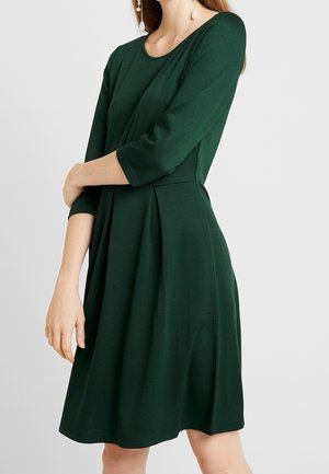 Femme portant une robe vert foncé mi-longue avec des manches trois-quarts et une jupe plissée, debout les bras croisés.