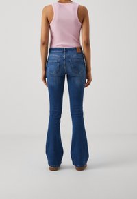 Jeans acampanados de color azul oscuro con cintura media, que presentan bolsillos traseros y un parche de cuero marrón en la cinturilla. La parte superior es una camiseta sin mangas rosa.