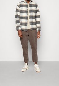 Veste en flanelle à carreaux grise et crème avec des accents beiges, fermeture boutonnée devant, poches poitrine, portée avec un pantalon marron fuselé et des baskets blanches.