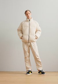 Beige pufferjakke med lynlås, der har en høj krave og lomme detaljer. Sammen med beige bukser og sorte sneakers med gule accenter.