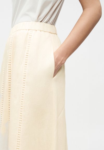 Γυναίκα που φοράει κρεμ φούστα με διακοσμητικές κατακόρυφες περικοπές και ελαστική μέση, χέρι τοποθετημένο στην δεξιά τσέπη, λευκό τοπ ορατό.