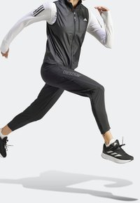 Czarna lekka sportowa kamizelka z zamkiem z przodu, zestawiona z czarnymi zwężanymi spodniami z nadrukiem "ADIZERO" i wąskim krojem, noszona z czarnymi tenisówkami.