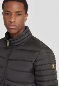 Zwart pufferjack met een hoge kraag, voorzien van horizontale stiksels en een logo patch op de linker schouder. Glad, lichtgewicht materiaal.