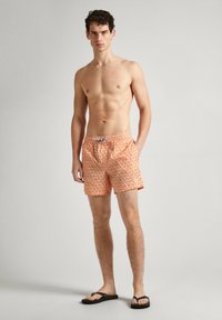 Oranžinės maudymosi šortai su balta lapų raštu, elastinga juosta ir raišteliu, derinami su juodais šlepetėmis. Lygi audinio tekstūra.