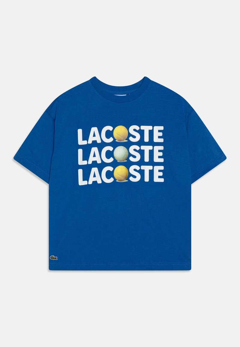 Camiseta azul de manga corta con "LACOSTE" repetido tres veces, cada vez con una pelota amarilla de tenis o una pelota blanca de golf reemplazando la letra "O".