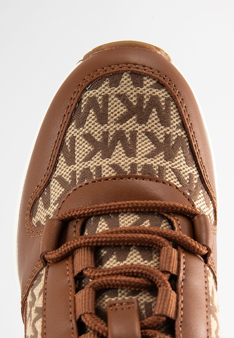 Zapatilla textil marrón con un patrón geométrico tejido y acentos en marrón oscuro, ojales de cuero marrón y una suela intermedia texturizada.