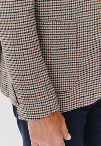 Brązowa marynarka w kratę houndstooth w odcieniach kremowych z niebieskimi i czerwonymi akcentami, teksturowany materiał i kieszeń z przodu.