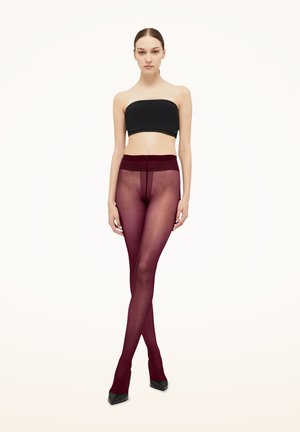 Vrouw staande, draagt een zwarte strapless top en doorschijnende bordeauxkleurige panty met zwarte puntige hakken tegen een effen lichte achtergrond.