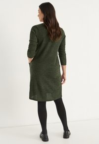 Knitklänning i mörkgrön, med trekvartsärmar och sidfickor. Tyget har en texturerad yta och en knälång klippning.
