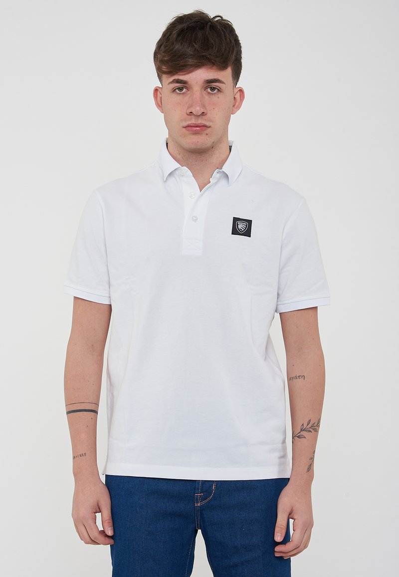 Polo bianco in cotone, con colletto pieghevole e patta con tre bottoni. Include una piccola patch nera con logo sul lato sinistro del petto.