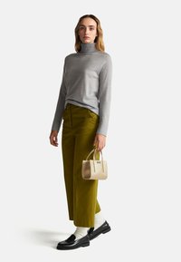 Maglione grigio a collo alto, pantaloni a gamba larga verde oliva, mocassini neri, calze bianche, mentre tiene una piccola borsa dorata. Design semplice, texture liscia.