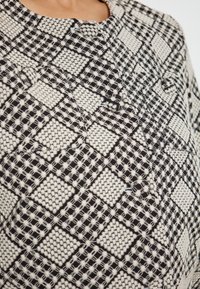 Maglione lavorato a maglia con un motivo geometrico in bianco e nero, caratterizzato da quadretti testurizzati e dettagli di cucitura evidenti. Collo rotondo e decorazione con bottoni.