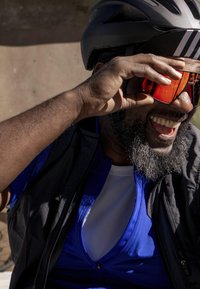 Casco nero con strisce bianche, occhiali da sole rossi, gilet nero sopra una maglietta da ciclismo blu. La persona si sta asciugando il sudore dalla fronte e sorride.