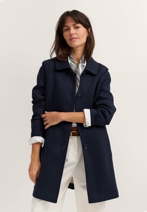 Frau trägt einen marineblauen Mantel über einem weißen Hemd mit aufgekrempelten Ärmeln und weißen Hosen, steht vor einem schlichten hellen Hintergrund.