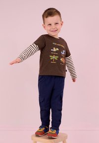 Brun grafik T-shirt med randiga långärmar, mörkblå cargobyxor och flerfärgade sneakers. Barnet poserar med en arm utsträckt.