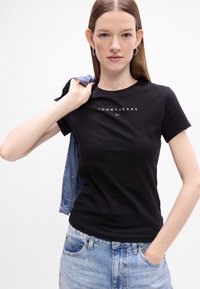 Melna kokvilnas T-krekls ar centrālo "TOMMY JEANS" logo baltā krāsā, apvienots ar gaiši ziliem džinsu šortiem, uz kuriem uzlikta gaiši zila jakas mala, kas karājas pāri vienai plecam.