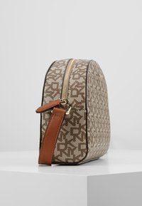 Pyöreämuotoinen ruskea kuvioitu laukku, jossa on teksturoitu kangas, kultainen vetoketju ja ruskea nahkahihna. Yksityiskohdissa hienovarainen brändin logo.
