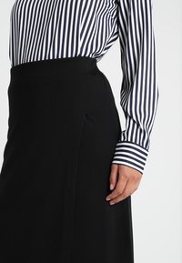 Jupe crayon noire avec fente sur le côté, associée à une chemise à rayures navy et blanche à manches longues. Le tissu semble lisse et structuré.