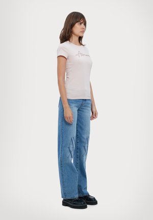Camiseta de algodón color rosa claro con logotipo en cursiva, combinada con jeans de mezclilla azul de pierna ancha que presentan un patrón de logotipo destacado en la parte frontal. Zapatos de plataforma negros.