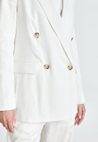 Blazer bianco doppiopetto con bottoni dorati, tessuto testurizzato, spalle strutturate e tasche laterali. Vestibilità su misura evidente.