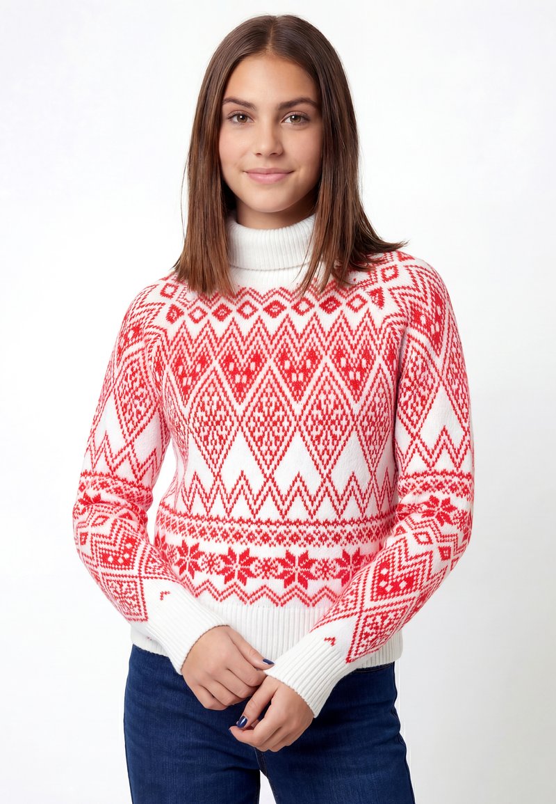 Joven con cabello castaño liso que lleva un suéter blanco de cuello alto con patrones geométricos y florales rojos, de pie frente a un fondo blanco simple.