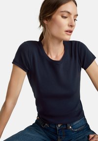 Tmavě modré přiléhavé tričko s krátkým rukávem vyrobené z měkké látky s kulatým výstřihem a žebrovaným lemem. Nosené s džíny.