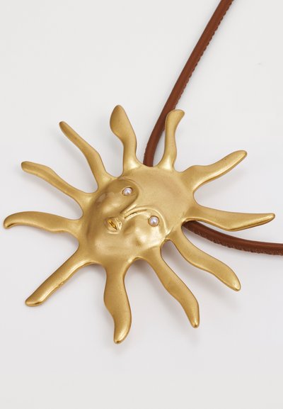 Goldener, sonnenförmiger Anhänger mit glatter Oberfläche, der zwei kleine Perlen als Augen hat und an einem braunen Lederband hängt.