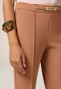 Pantalons couleur pêche avec une texture lisse, ornés d'une chaîne en or à la taille. Un bracelet manchette en or décore le poignet.